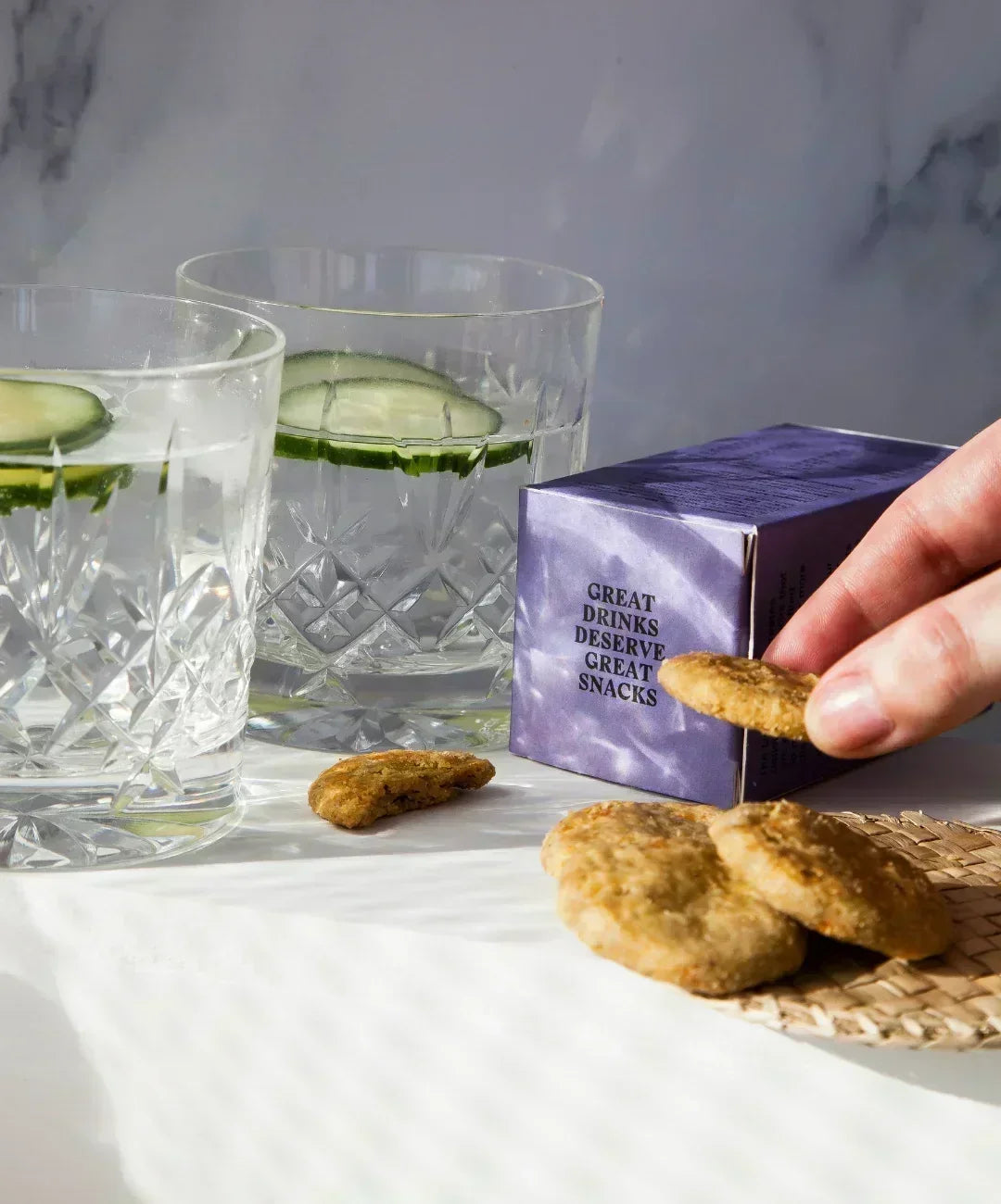 Two crystal glasses with Gin and Tonic with cucumber garnish, a purple box of Drinks Biscuits, several savoury biscuits in the foreground with someones hand picking one up.