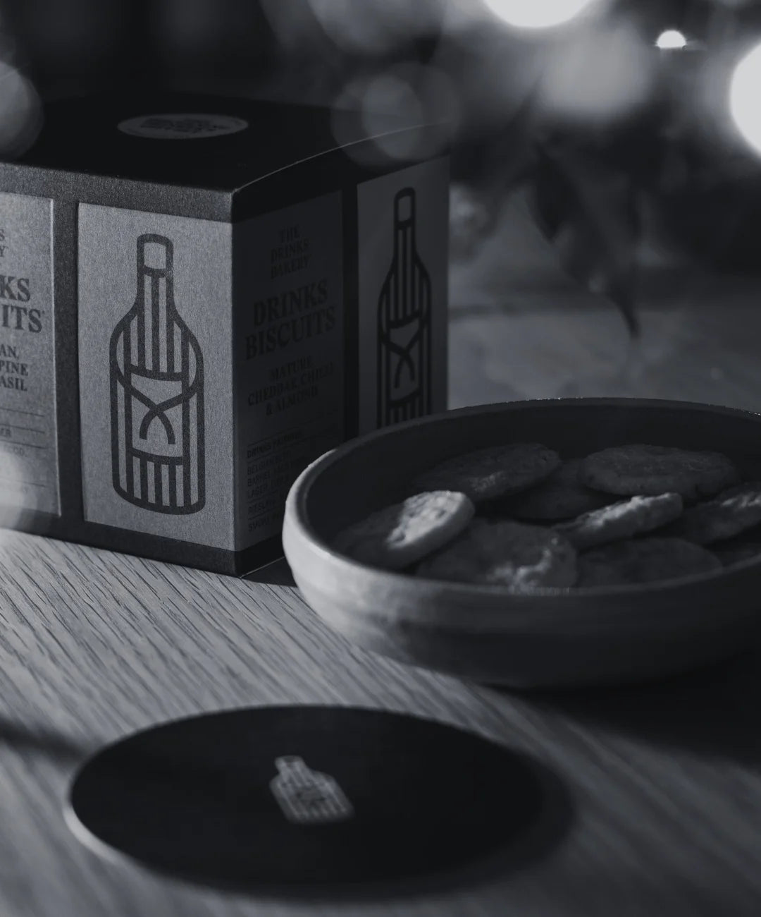 A dark grey and white box of Drinks Biscuits gift box with a bottle illustration, shown next to a bowl of savoury biscuits on a wooden surface with a blurred background.