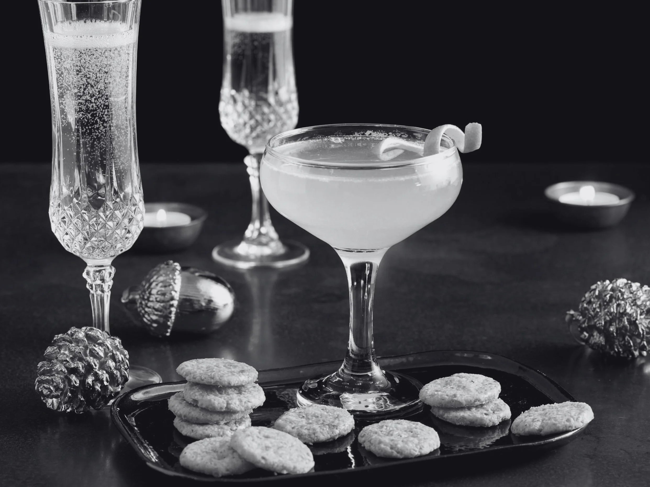 A black-and-white styled scene with a coupe glass filled with a pale drink, two tall champagne flutes, a tray of small round savoury biscuits, pinecones, and a dark background with candles.