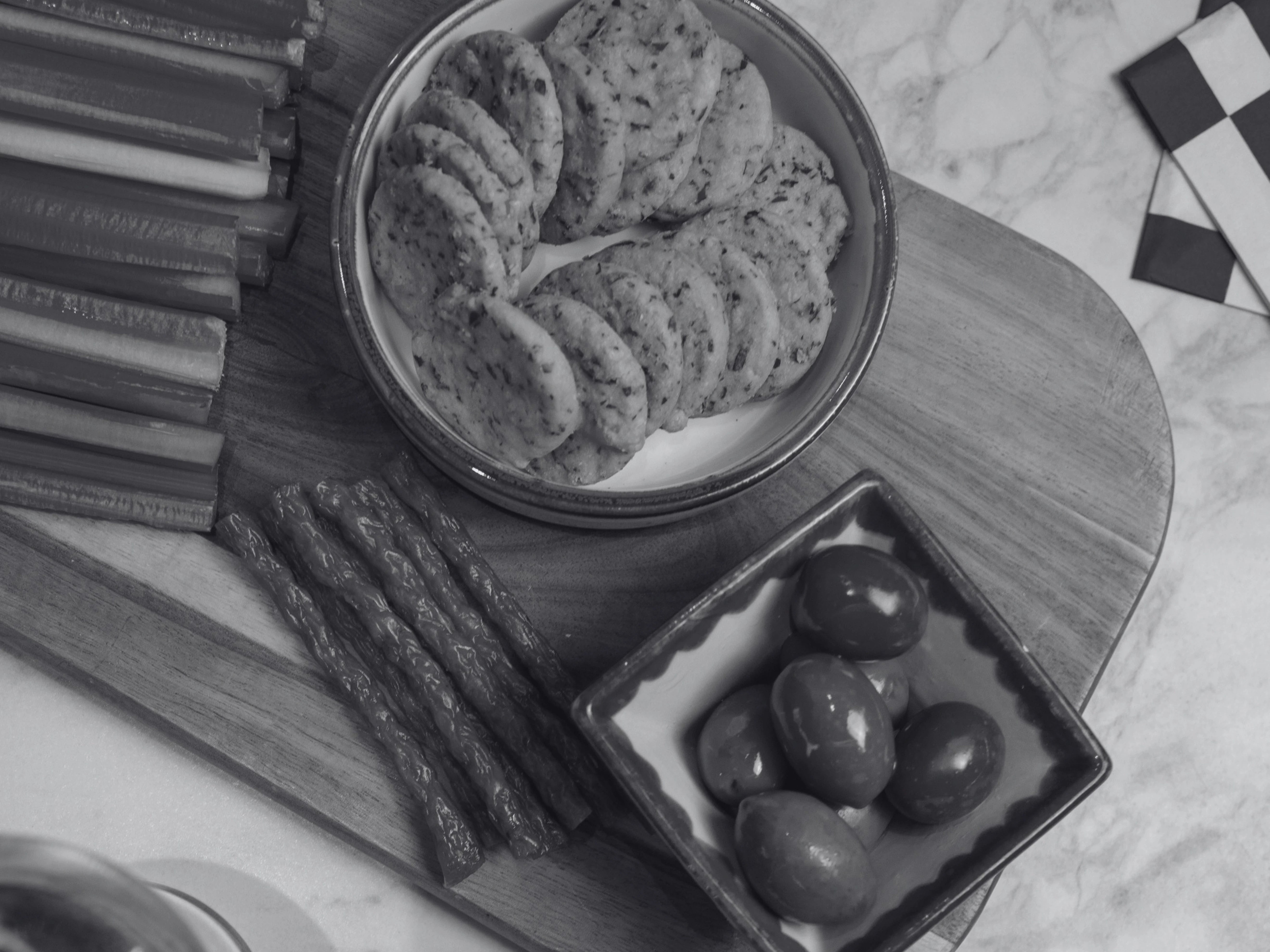 A styled black-and-white food scene showing a bowl of round savoury biscuits, several long celery sticks and thin sausage snacks, and a small dish of olives arranged on a wooden board over a marble surface.