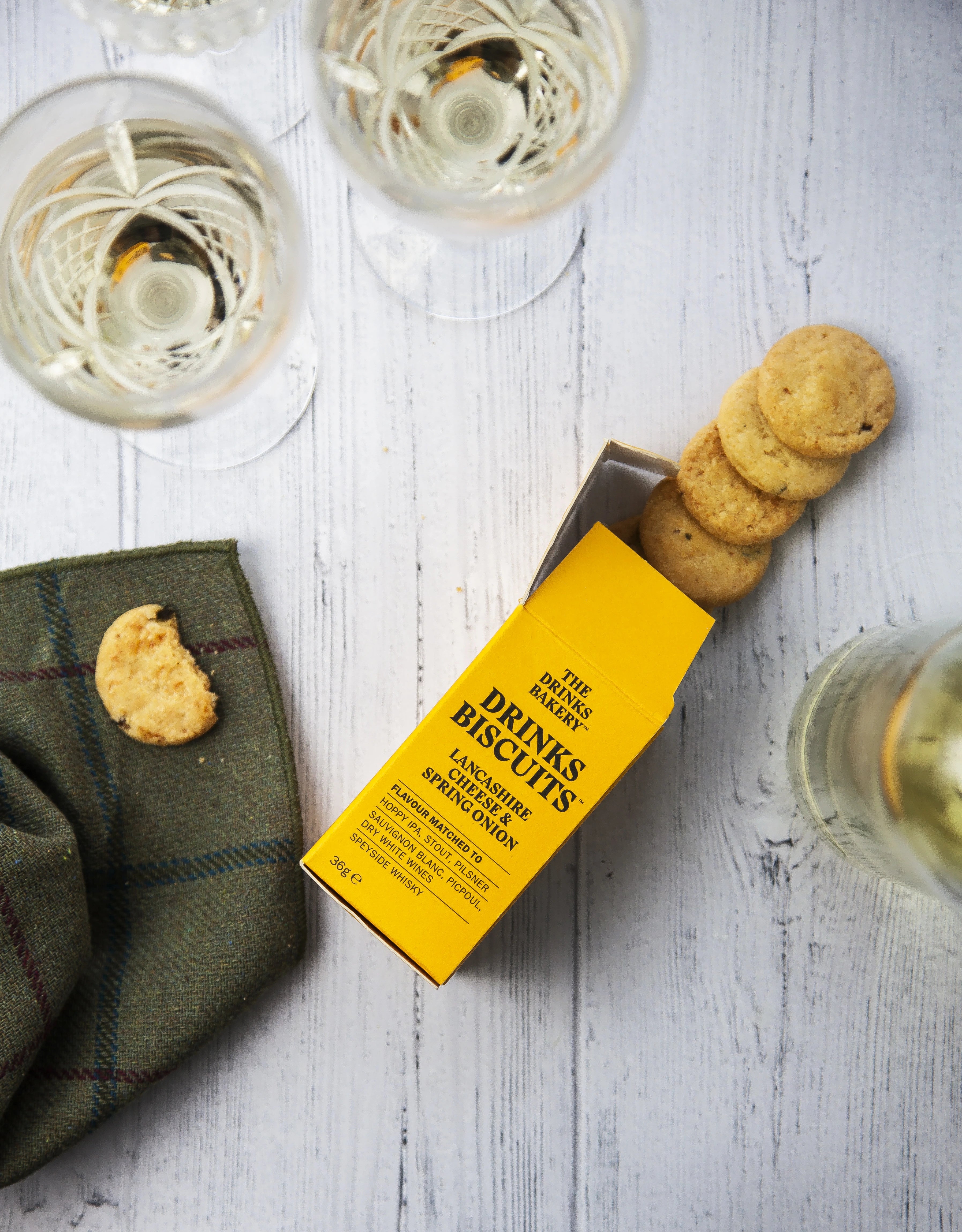 A yellow box of Drinks Biscuits lies open on a white wood surface, with several round biscuits stacked out of the box and a few biscuits on a green cloth. Two wine glasses are partially visible around the scene.