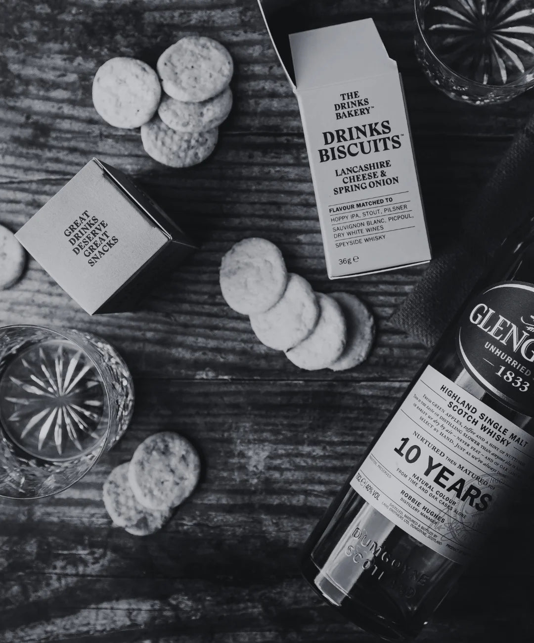 Bottle of Glenlivet 10 Year Old whisky, 'The Drinks Bakery' biscuits, and glasses on a wooden surface.