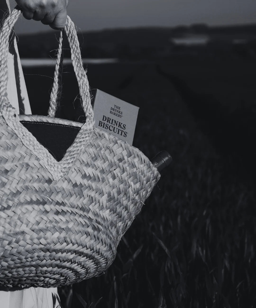 Woven tote bag held by a person with a box of savoury 'Drinks Biscuits' and bottle of wine sticking out of it. On a field of grass.