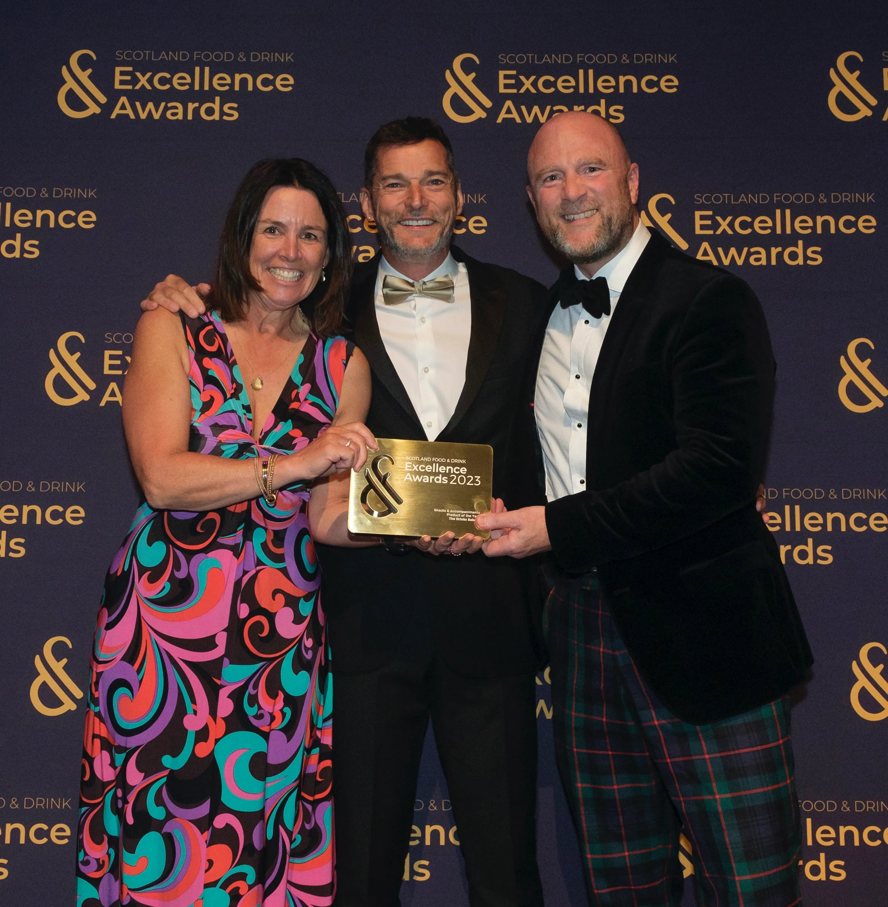 Three people standing together on a dark blue Scotland Food & Drink Excellence Awards backdrop, holding a gold award plaque. Sarah Fraser (the woman) wears a colourful abstract print dress,  Fred Sirieix (the man in the centre) wears a black tuxedo with a gold bow tie, and Andy Murray (the man on the right) wears a black tuxedo jacket with a black bow tie and tartan kilt.