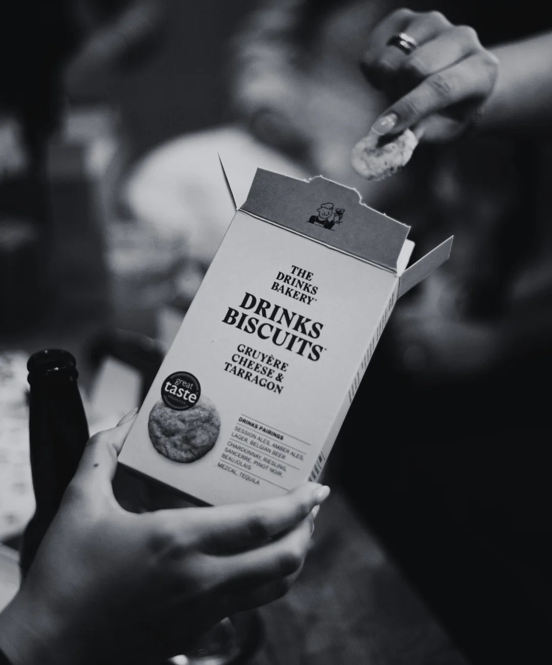 A person holds a box of The Drinks Bakery Drinks Biscuits Gruyere Cheese and Tarragon in a dimly lit setting, with another hand reaching in from the upper right to get one of the biscuits.