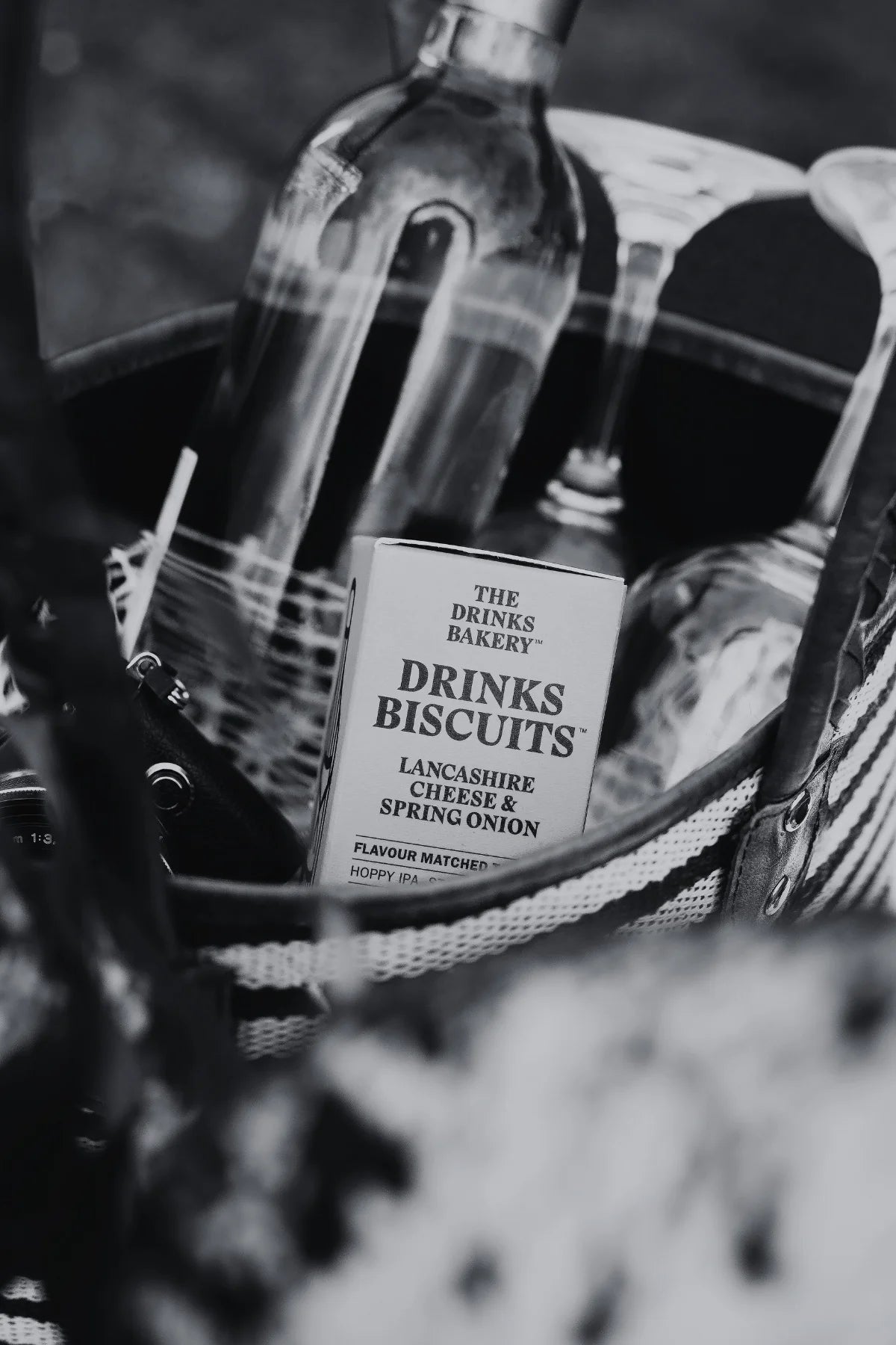 A black and white close-up of a woven bag containing a box of Drinks Biscuits, with a glass bottle of wine and two glasses behind it.