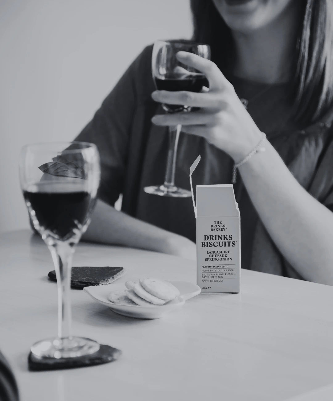 Person holding a glass of red wine with a box of Drinks Biscuits on a table. Drinks Biscuits are savoury cheese biscuits expertly flavour profiled to pair with drinks.