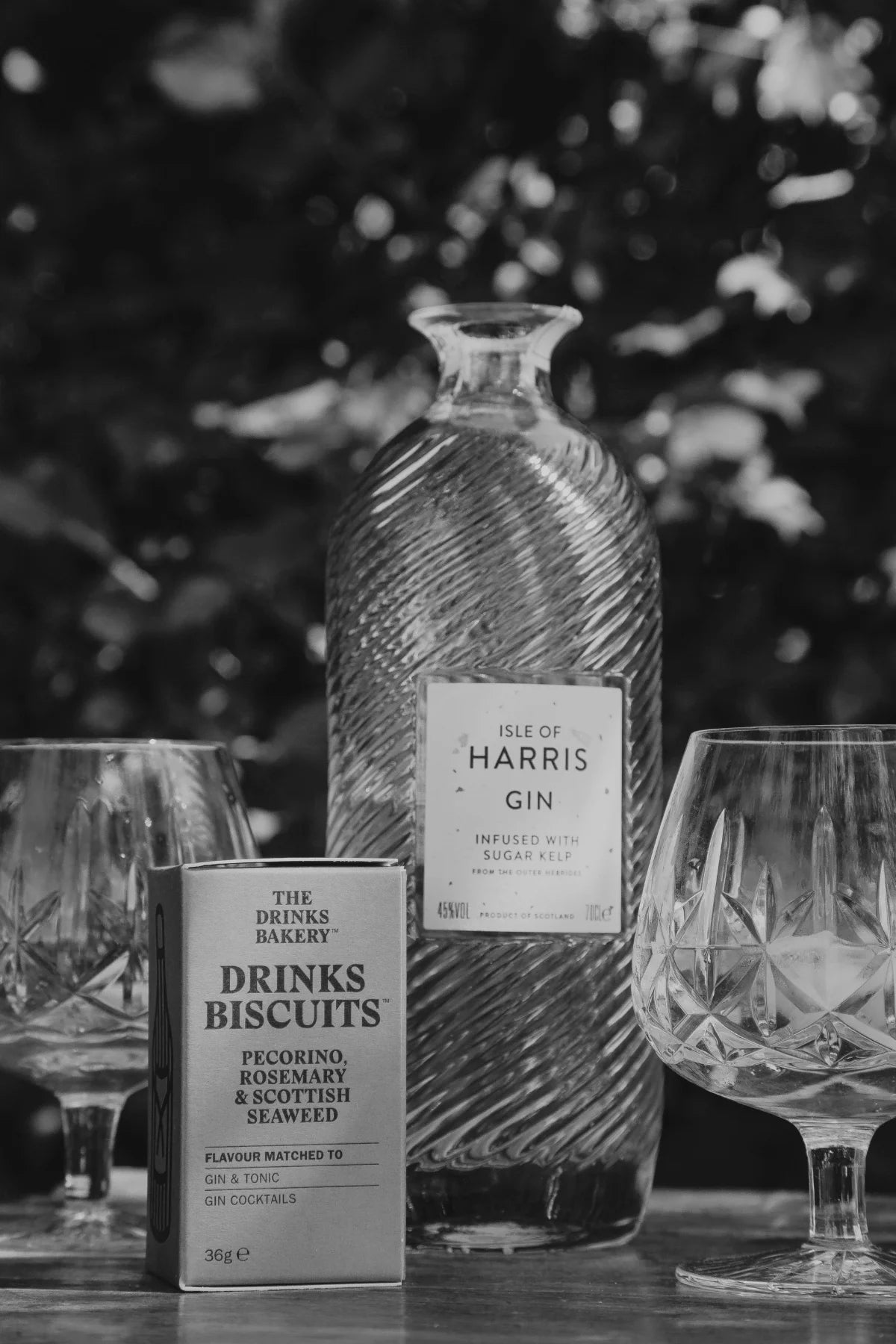 Black and white image including Pecorino Cheese & Rosemary Drinks Biscuits, Isle of Harris Gin, two crystal glasses with ice and gin and tonic, on a table. 