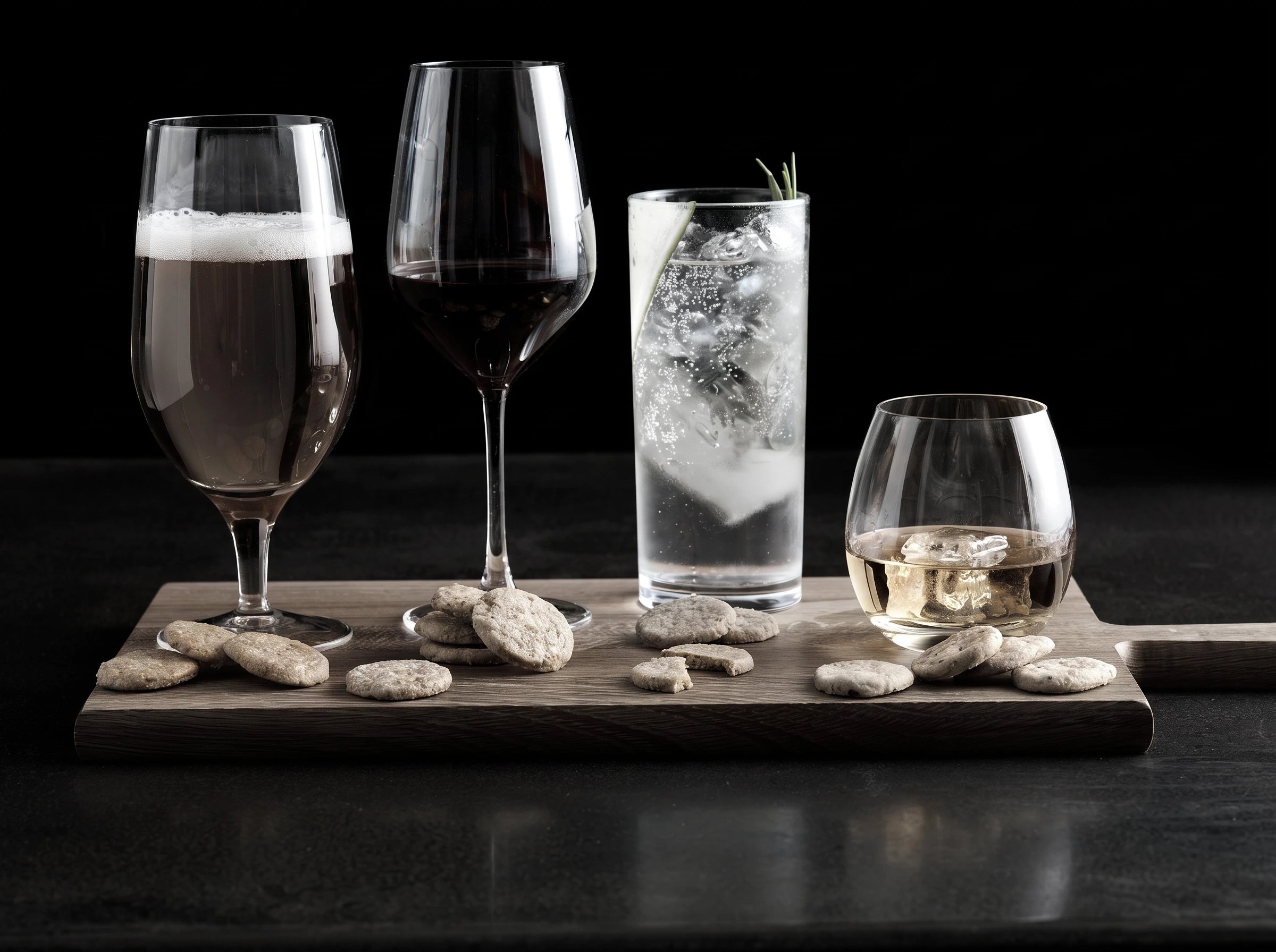 Four clear drinking glasses arranged on a wooden board with stones, set against a dark studio background. The glasses include a bubbly beer glass, a dark red wine glass, a tall clear glass with gin and tonic, ice and a sprig, and a small glass with a pale golden whisky.