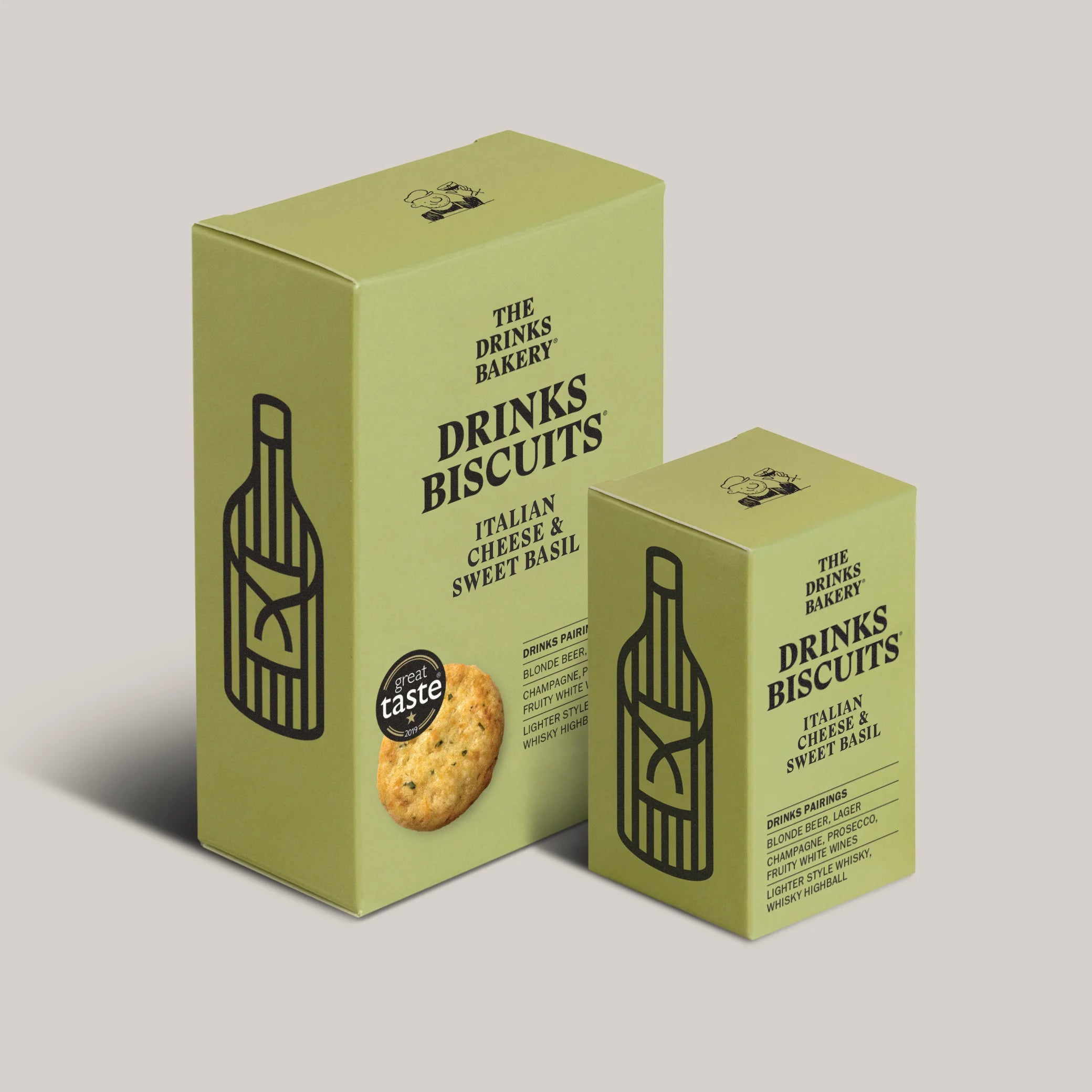 Two light olive green product boxes for 'The Drinks Bakery' 'Drinks Biscuits' shown on a solid light gray background. The larger box features a black line art illustration of a bottle, a photo of a biscuit, and a 'taste' badge, while the smaller box is positioned in front of it.