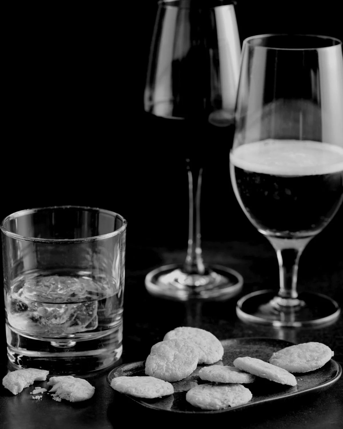 Black and white image of three glasses of alcoholic drinks, one with whisky, one with red wine and one with a beer. There is also a tray of savoury biscuits are the foreground of the image.