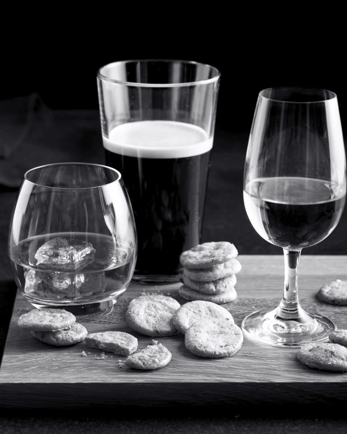 Three glasses on a wooden chopping board, one with whisky, one with a dark ale, and one with white wine. There are also savoury biscuits on the board.