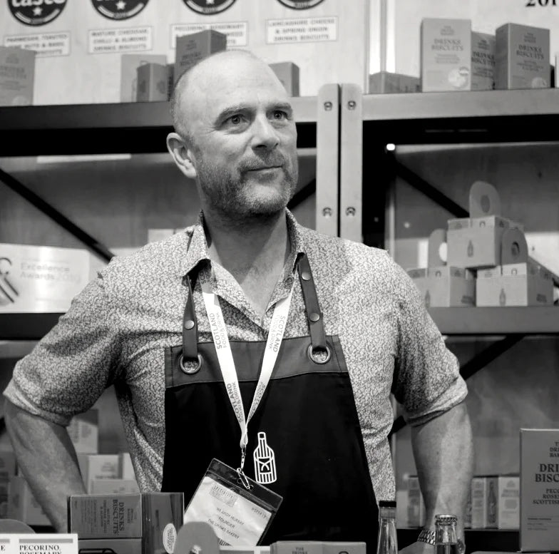 A black and white photo of a man wearing a patterned shirt and a dark apron with a hangtag, standing behind a counter with shelves of product boxes in the background.