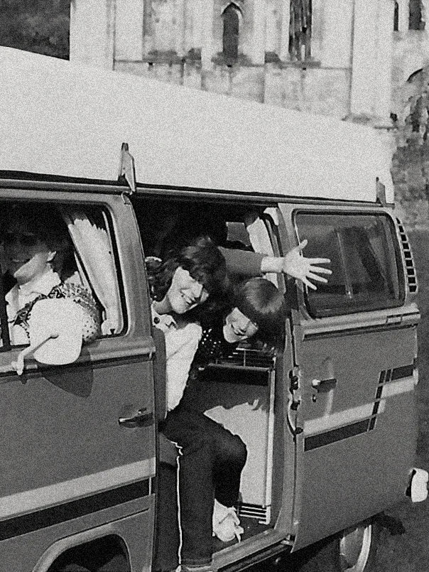 Black and white photograph of three people sitting inside and beside a vintage camper van outdoors, with a stone building in the background. I.e. Andy (Founder of The Drinks Bakery) in his family camper van where it all began.