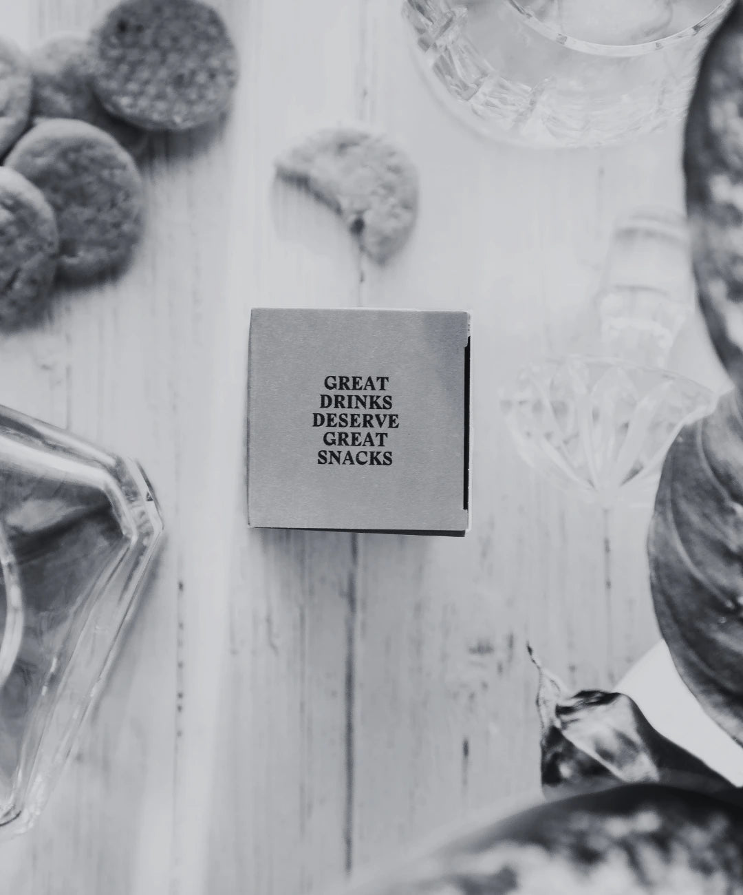 Small square box of savoury biscuits with 'GREAT DRINKS DESERVE GREAT SNACKS' text on a wooden surface with snacks around. Whisky decanter in the corner.